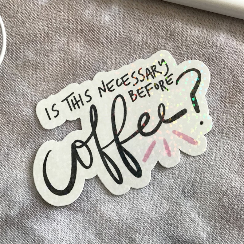 May include: A white sticker with the text "IS THIS NECESSARY BEFORE coffee?" in black script. The sticker has a scalloped edge and holographic accents, with pink and red bursts emanating from the word "coffee".