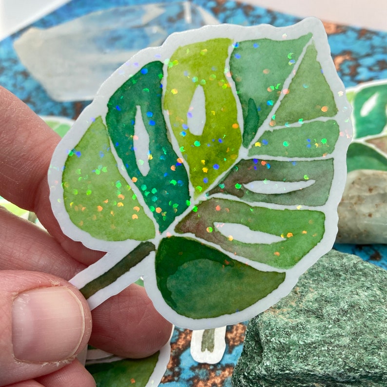 K&ouml;nnte beinhalten: Ein Aquarell-Aufkleber eines Monstera-Blattes, mit Gr&uuml;nt&ouml;nen und braunen und schillernden Glitzerakzenten. Das Blatt hat einen wei&szlig;en Rand. Der Aufkleber wird vor einem strukturierten Hintergrund gehalten.