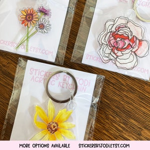 Puede incluir: Llaveros acr&iacute;licos con dise&ntilde;os florales. Uno muestra un ramo de flores naranjas, rosas y blancas. Otro muestra una rosa rosa y roja. Un tercer llavero tiene un girasol amarillo. El texto "STICKERS BY TODI ACRYLIC KEYCHAIN" es visible.