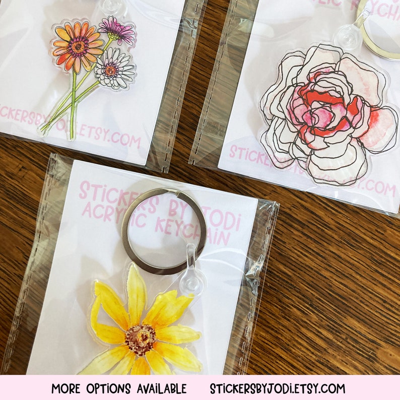 Puede incluir: Llaveros acr&iacute;licos con dise&ntilde;os florales. Uno muestra un ramo de flores naranjas, rosas y blancas. Otro muestra una rosa rosa y roja. Un tercer llavero tiene un girasol amarillo. El texto "STICKERS BY TODI ACRYLIC KEYCHAIN" es visible.