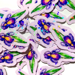 May include: A collection of stickers featuring watercolor iris flowers. The stickers have a light purple background with a white border. The irises are purple and yellow with green leaves. Holographic glitter accents the stickers.