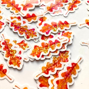May include: A collection of autumn-themed stickers featuring oak leaves. The stickers have a white border and are in shades of orange, red, and yellow, with iridescent glitter accents. The leaves are detailed with veins and textures.