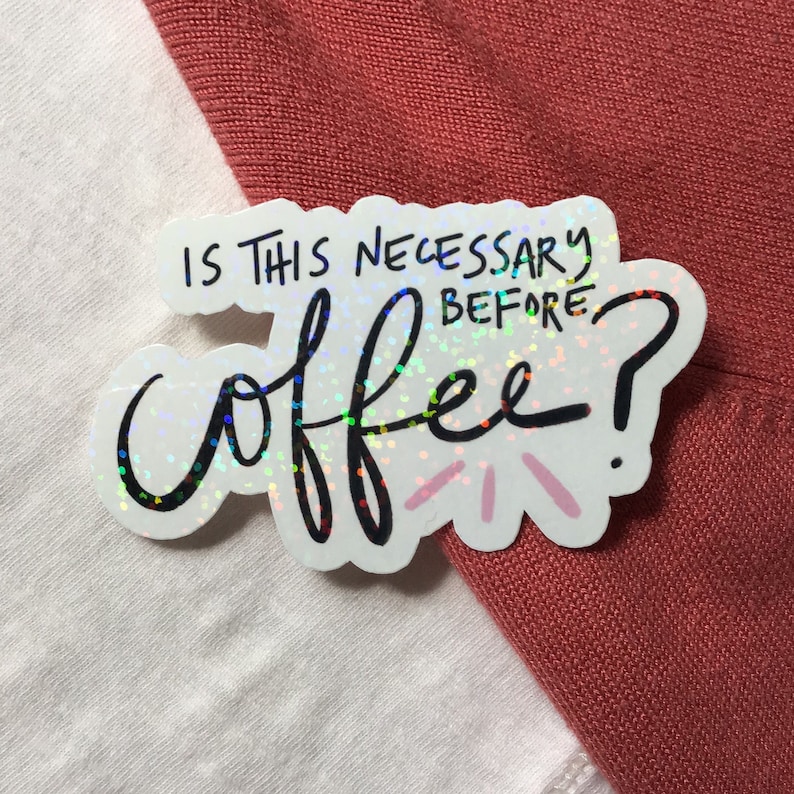 May include: A holographic sticker with the text "IS THIS NECESSARY BEFORE coffee?" in black script. The sticker is white with a colorful, iridescent sheen and is placed on a white and red-orange fabric background.