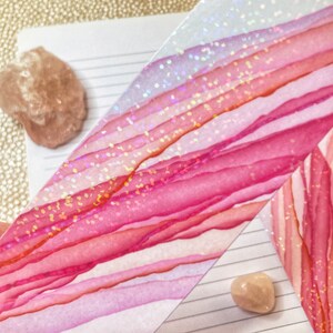 May include: Close-up of a watercolor design with shades of pink and red, accented with glitter. The design is on a white background with lines, and there are two small, light-colored stones.