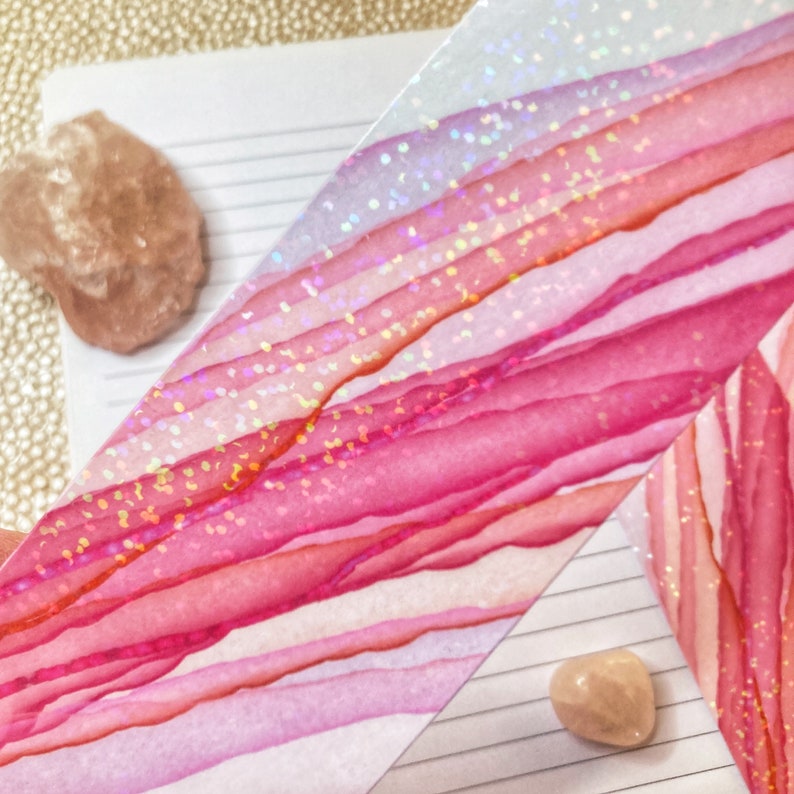 May include: Close-up of a watercolor design with shades of pink and red, accented with glitter. The design is on a white background with lines, and there are two small, light-colored stones.
