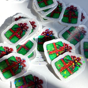 May include: A collection of stickers featuring a green gift box design with a red bow. The stickers have a white border and a glittery, holographic effect. The gifts are scattered across a white surface.