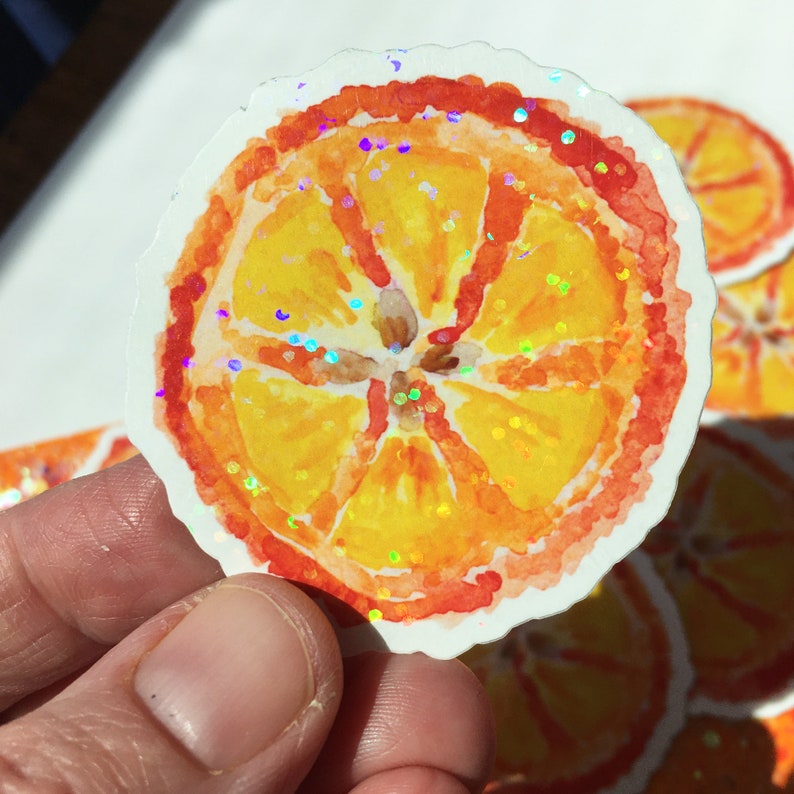 Puede incluir: Una pegatina de acuarela de una rodaja de naranja con borde blanco. La rodaja de naranja es amarilla con segmentos naranjas y una c&aacute;scara roja. La pegatina tiene detalles de purpurina iridiscente.