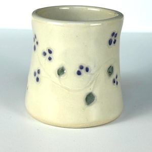 May include: A small, white ceramic cup with a floral design. The cup has a vine pattern with green leaves and purple berries.