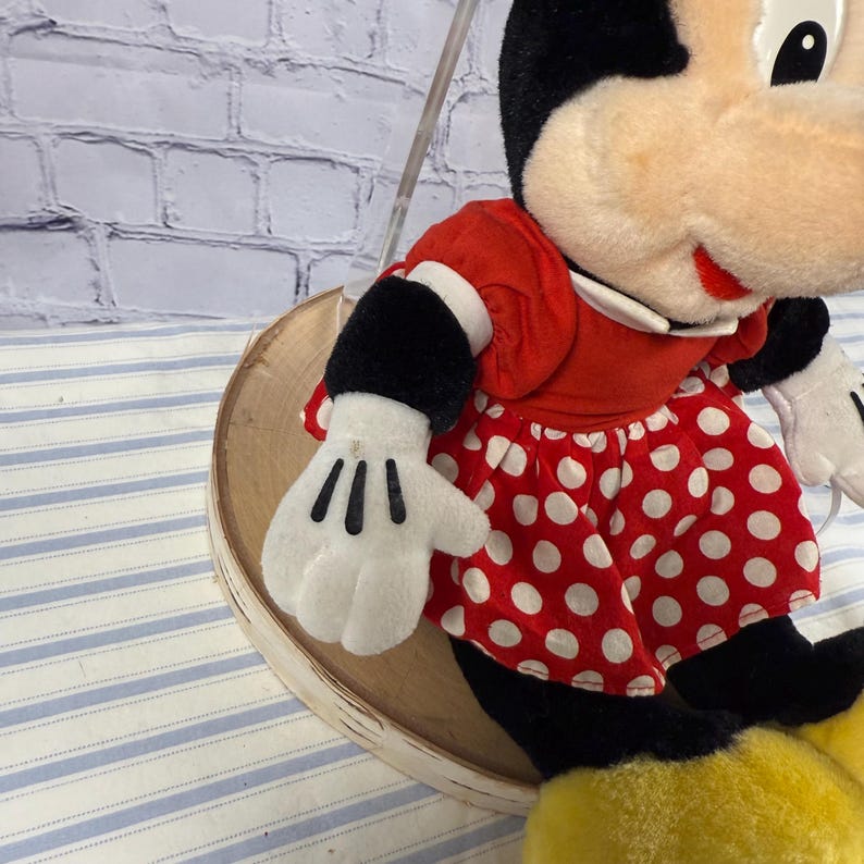 May include: A plush Minnie Mouse doll, featuring a red dress with white polka dots, black gloves with three black lines, and black shoes. The doll has a white face, black ears, and a red bow. The doll is on a wooden display.
