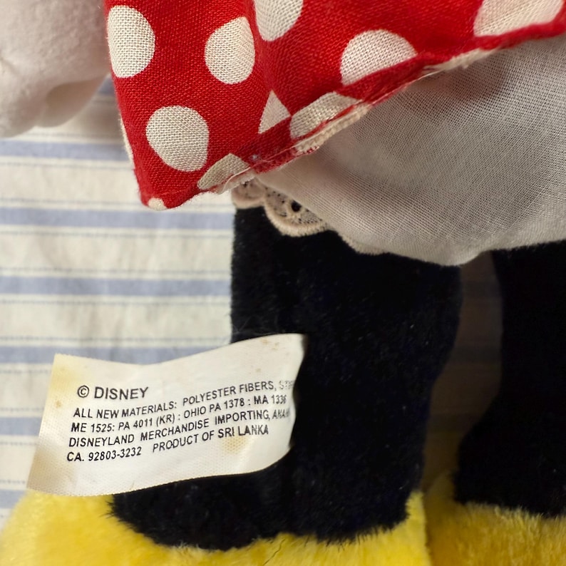 May include: A plush toy featuring a red dress with white polka dots, a white underskirt, and black legs. The toy has yellow feet and a tag with the Disney logo and product information.