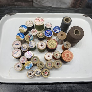 May include: A collection of vintage sewing thread spools in various sizes and colors, including brands like Coats & Clark, Star, and Lily. The spools feature paper labels with printed text and numbers, arranged on a white rectangular metal tray.