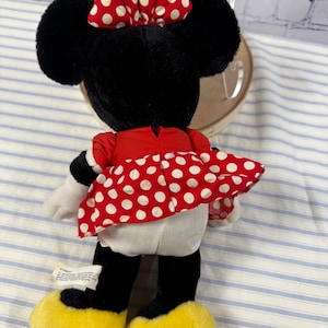 May include: A plush Minnie Mouse doll, viewed from the back, features black fur, large ears, and a red dress with white polka dots. The doll has a red and white polka dot bow and yellow shoes. A Disney tag is visible.