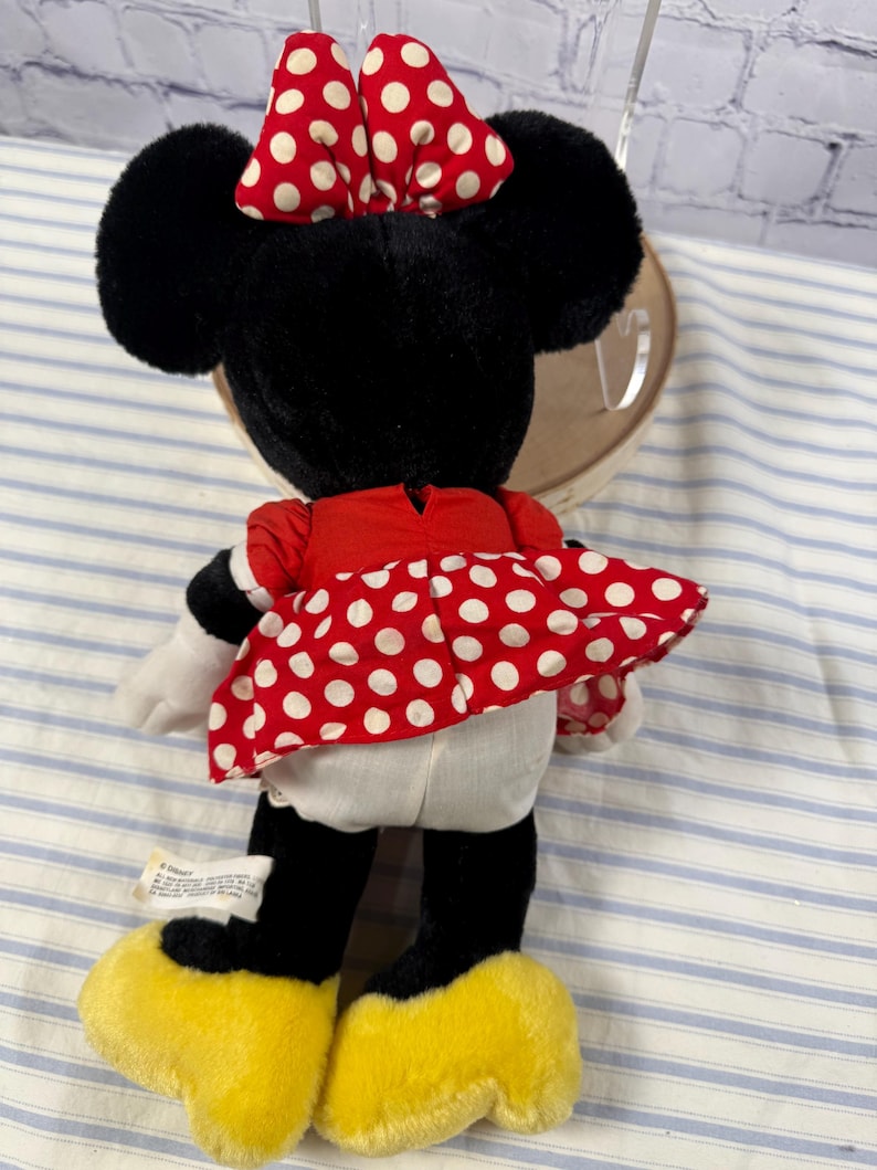 May include: A plush Minnie Mouse doll, viewed from the back, features black fur, large ears, and a red dress with white polka dots. The doll has a red and white polka dot bow and yellow shoes. A Disney tag is visible.