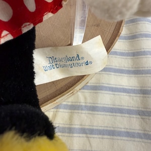 May include: A close-up of a plush toy with a black and yellow body, a red and white polka dot bow, and a tag that reads "Disneyland&reg; Walt Disney World&reg;". The tag is attached to a wooden hoop.