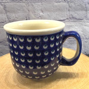 May include: A ceramic mug with a dark blue exterior adorned with a repeating pattern of white and blue crescent shapes. The interior and rim are cream-colored, and the handle is a matching dark blue. The mug sits on a wooden surface.