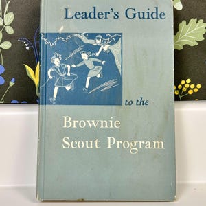 May include: A blue book with the title "Leader's Guide to the Brownie Scout Program". The cover features a black and white illustration of three girls in uniform playing outdoors.