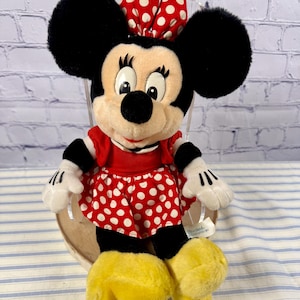 May include: A plush Minnie Mouse doll, featuring large black ears, a red and white polka dot bow, and a matching skirt. The doll has a red top, white gloves, and yellow shoes. The background is a brick wall and a blue and white striped surface.