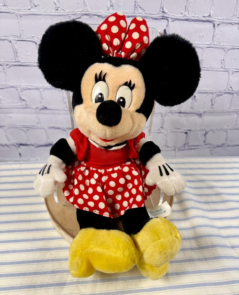 May include: A plush Minnie Mouse doll, featuring large black ears, a red and white polka dot bow, and a matching skirt. The doll has a red top, white gloves, and yellow shoes. The background is a brick wall and a blue and white striped surface.