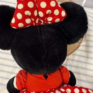May include: A plush Minnie Mouse doll, viewed from the back, featuring black ears and a red bow with white polka dots. The doll wears a red dress with white polka dots. The background is a blue and white striped fabric.