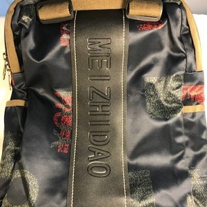 May include: A black and blue patterned backpack with a black leather strap that says "ME I ZHI DAO" in white lettering.