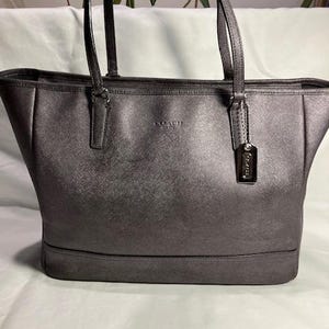 May include: A metallic gray tote bag with two top handles. The bag has a rectangular shape with a structured base and a subtle sheen. The word "COACH" is embossed on the front, and a small tag hangs from one handle.