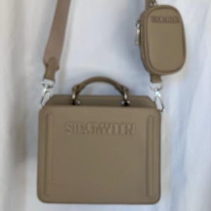 May include: A beige handbag with a matching small pouch. The handbag has a rectangular shape with a top handle and a long shoulder strap. The brand name "STEVE MADDEN" is embossed on the front of the handbag. The small pouch is attached to the strap.