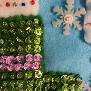 May include: Close-up of a felt craft with a light blue background. The craft features a green, sequined section, a white section with colorful beads, and a snowflake embellishment. The sequins are green and pink.