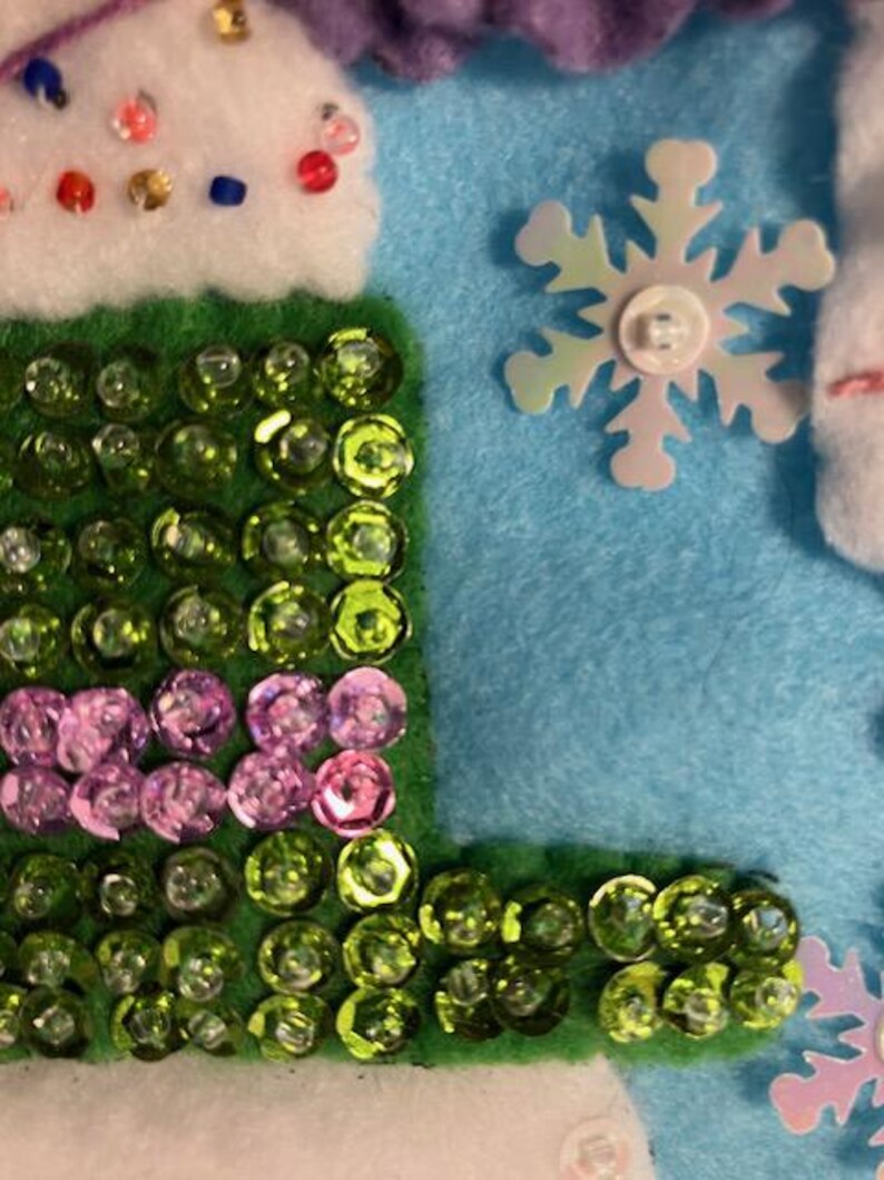 May include: Close-up of a felt craft with a light blue background. The craft features a green, sequined section, a white section with colorful beads, and a snowflake embellishment. The sequins are green and pink.