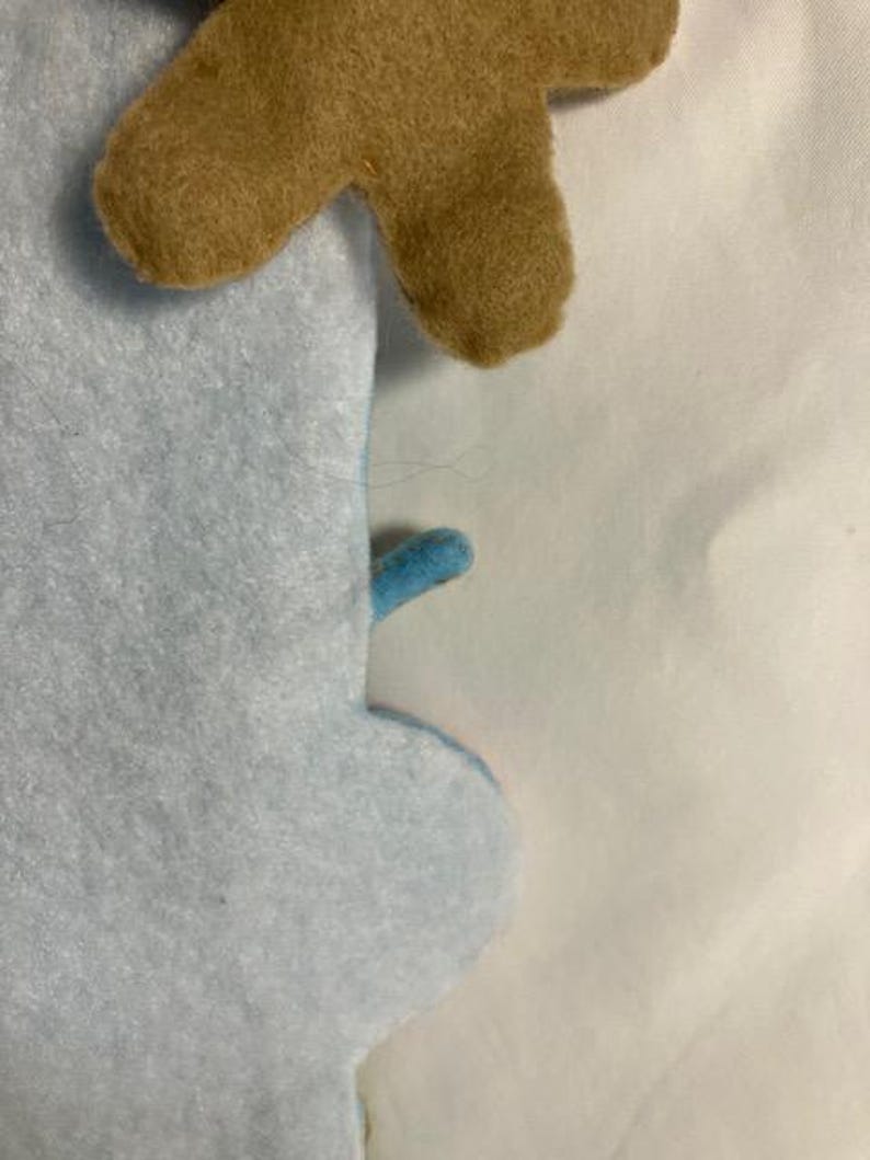 May include: Close-up of a light blue felt snowman with a brown felt antler. The snowman has a small blue felt arm. The felt is soft and textured.