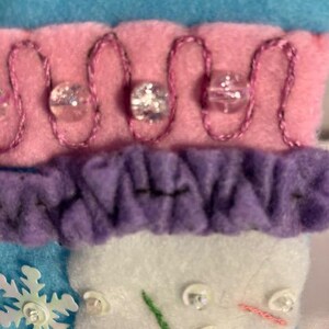 May include: Close-up of a felt Christmas stocking with a snowflake, beads, and embroidered details. The stocking features blue, pink, purple, and white sections. The pink section has a decorative stitch with clear beads.