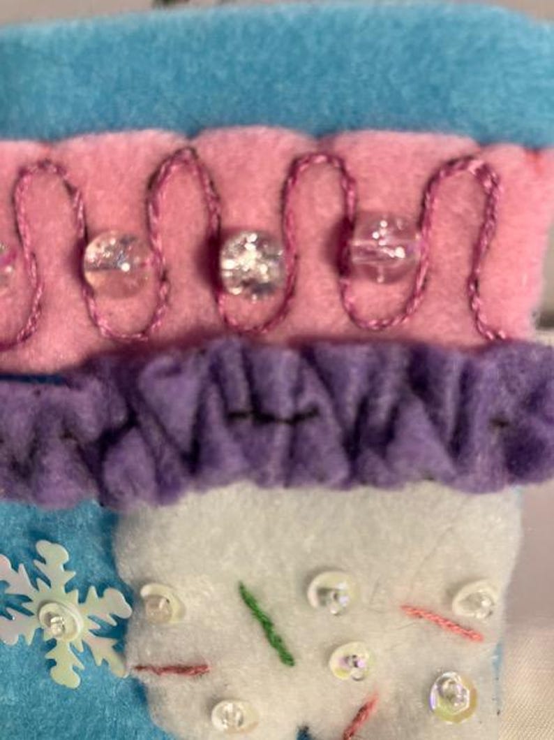 May include: Close-up of a felt Christmas stocking with a snowflake, beads, and embroidered details. The stocking features blue, pink, purple, and white sections. The pink section has a decorative stitch with clear beads.