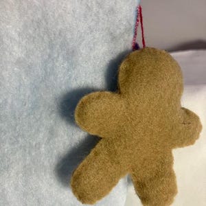 May include: A felt gingerbread man ornament in shades of brown, suspended by a red string. The ornament is attached to a light blue felt surface, suggesting a holiday theme. The gingerbread man has a simple, classic design.