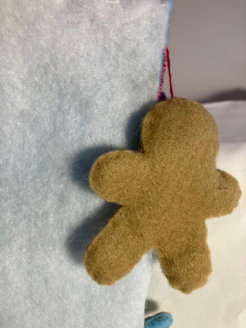 May include: A felt gingerbread man ornament in shades of brown, suspended by a red string. The ornament is attached to a light blue felt surface, suggesting a holiday theme. The gingerbread man has a simple, classic design.
