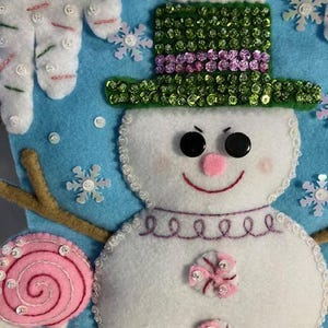 May include: A handmade Christmas stocking featuring a snowman design. The snowman has a green sequined hat, black button eyes, and a pink nose and mouth. The stocking is decorated with snowflakes, a pink lollipop, and gingerbread men.