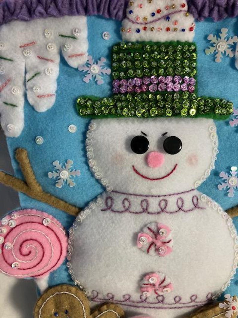 May include: A handmade Christmas stocking featuring a snowman design. The snowman has a green sequined hat, black button eyes, and a pink nose and mouth. The stocking is decorated with snowflakes, a pink lollipop, and gingerbread men.