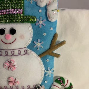 May include: A handmade Christmas stocking featuring a snowman design. The snowman has a green sequined hat, black eyes, and a pink nose. The stocking is decorated with snowflakes, candy canes, and gingerbread men.