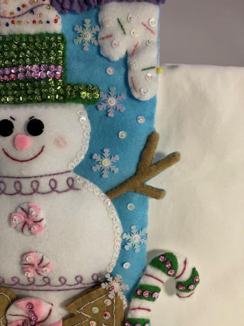 May include: A handmade Christmas stocking featuring a snowman design. The snowman has a green sequined hat, black eyes, and a pink nose. The stocking is decorated with snowflakes, candy canes, and gingerbread men.