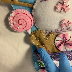 May include: A close-up of a felt Christmas stocking with candy and gingerbread man decorations. The stocking features a light blue background with a white snowflake and a brown branch. Pink and white candies and a gingerbread man are sewn onto the stocking.