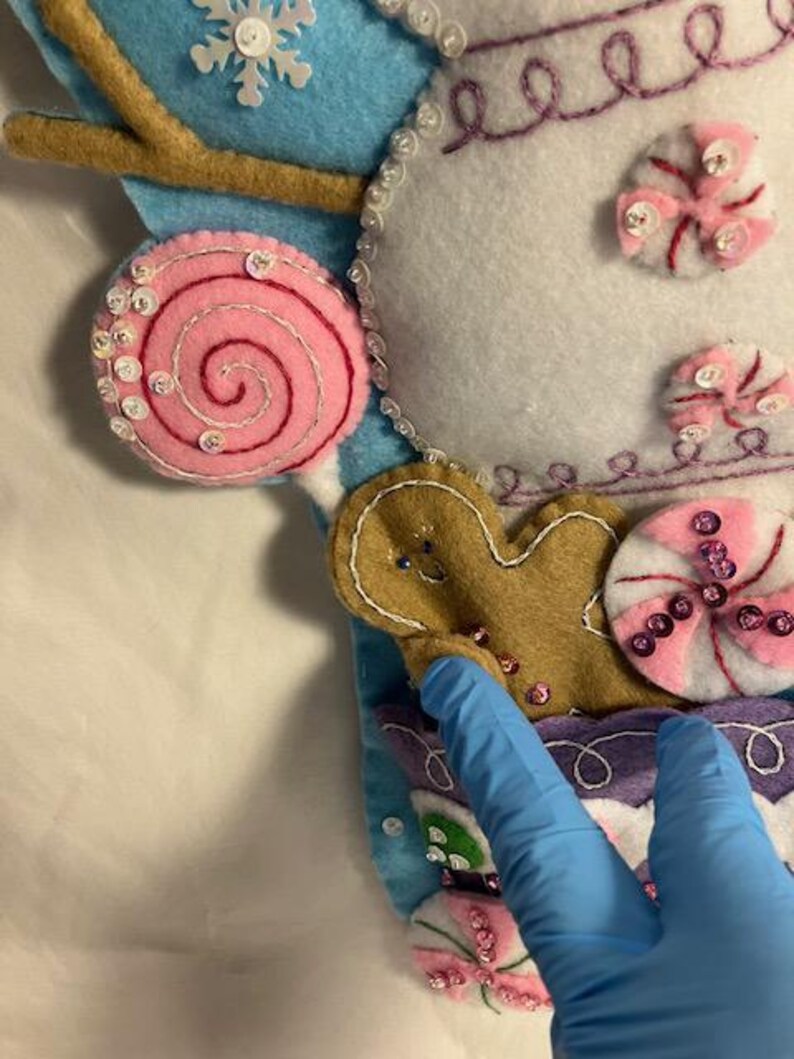 May include: A close-up of a felt Christmas stocking with candy and gingerbread man decorations. The stocking features a light blue background with a white snowflake and a brown branch. Pink and white candies and a gingerbread man are sewn onto the stocking.