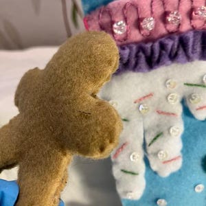 May include: A brown felt gingerbread man is held in front of a blue and white felt Christmas stocking. The stocking has pink and purple accents, with decorative sequins and bead embellishments. The stocking has a snowflake design.