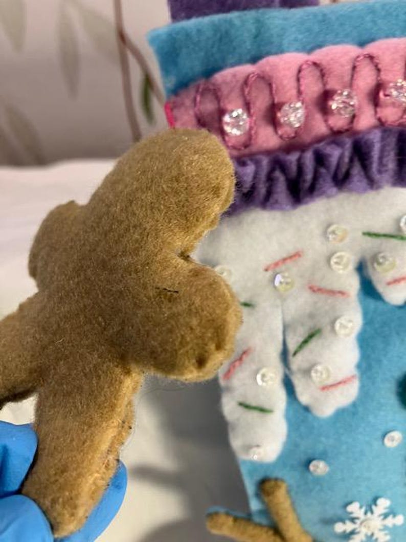 May include: A brown felt gingerbread man is held in front of a blue and white felt Christmas stocking. The stocking has pink and purple accents, with decorative sequins and bead embellishments. The stocking has a snowflake design.