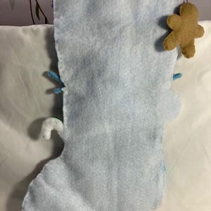 May include: A light blue felt Christmas stocking with a gingerbread man ornament. The stocking has a white candy cane and blue accents. The stocking is made of soft felt material.