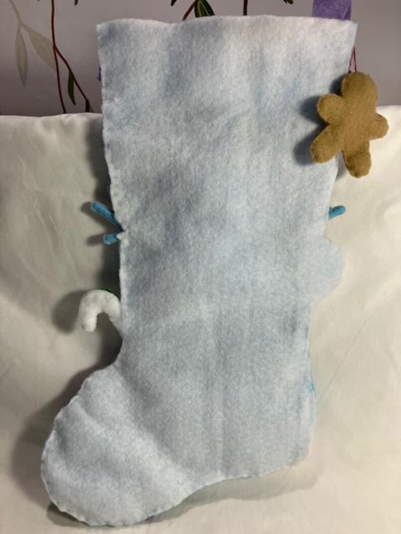 May include: A light blue felt Christmas stocking with a gingerbread man ornament. The stocking has a white candy cane and blue accents. The stocking is made of soft felt material.