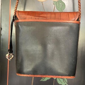May include: A black leather crossbody bag with a brown leather flap and a heart-shaped charm. The bag has a braided strap and a brown leather trim.