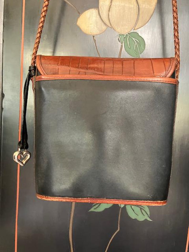 May include: A black leather crossbody bag with a brown leather flap and a heart-shaped charm. The bag has a braided strap and a brown leather trim.
