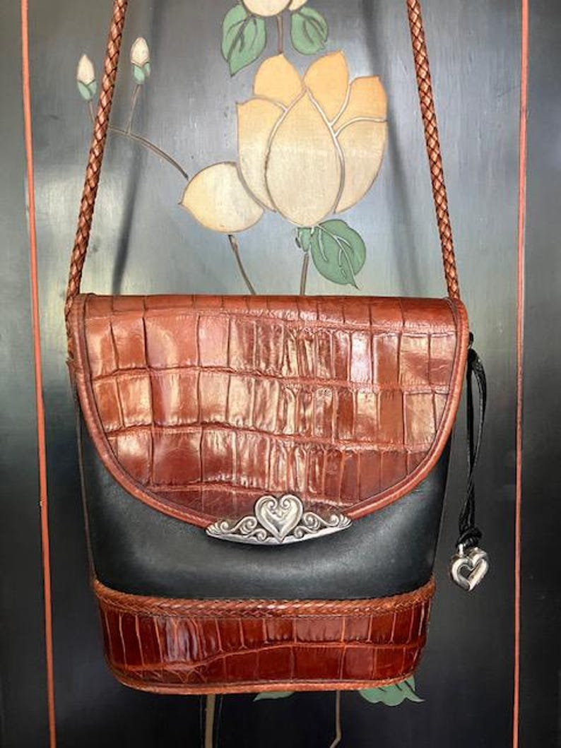 May include: A brown and black leather shoulder bag with a silver heart-shaped clasp. The bag has a braided strap and a small charm hanging from it.