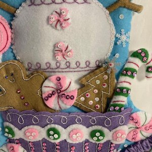 May include: A handmade Christmas stocking featuring a snowman design. The stocking is made of felt and decorated with gingerbread men, candy canes, and other festive elements. The color palette includes white, blue, pink, green, and brown.