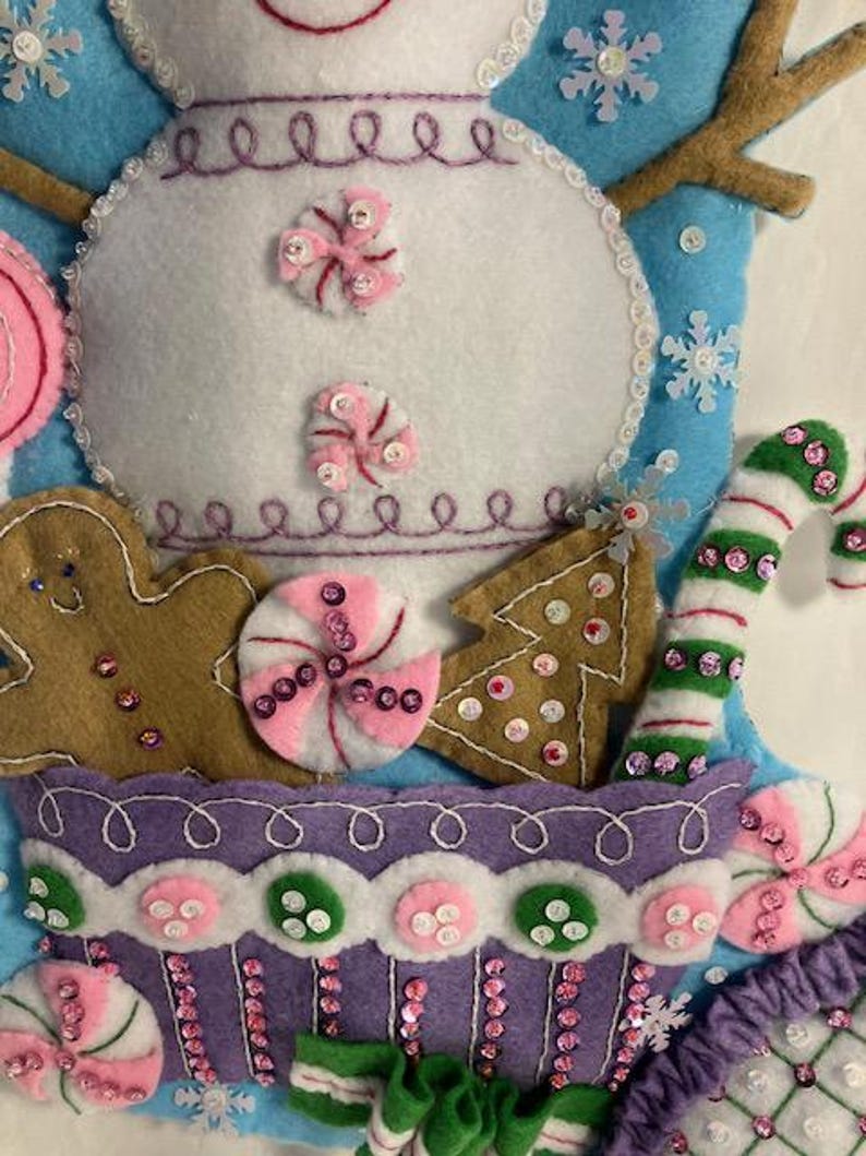 May include: A handmade Christmas stocking featuring a snowman design. The stocking is made of felt and decorated with gingerbread men, candy canes, and other festive elements. The color palette includes white, blue, pink, green, and brown.