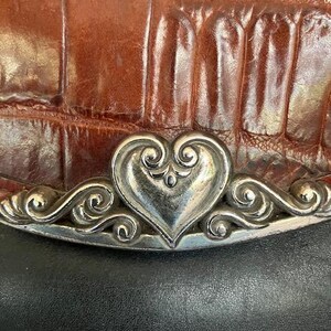 May include: A brown leather purse with a silver heart-shaped clasp. The clasp has intricate details and is surrounded by a silver border.