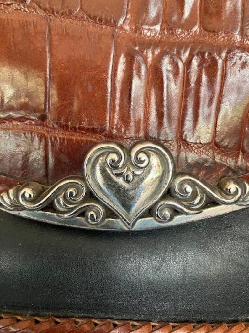 May include: A brown leather purse with a silver heart-shaped clasp. The clasp has intricate details and is surrounded by a silver border.